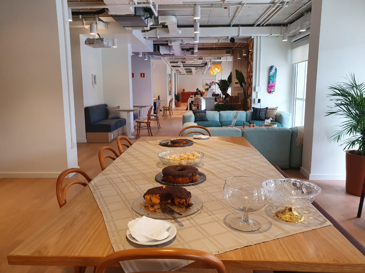 Mesa de jantar comprida de madeira com pratos de vidro em um lounge de escritório moderno e luminoso.