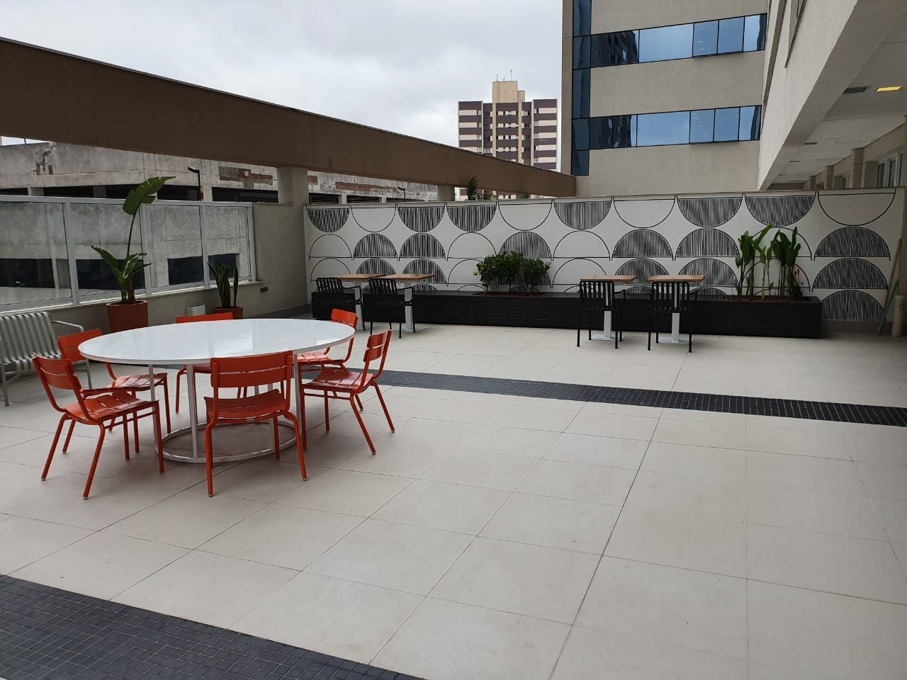 Pátio externo com cadeiras e mesas vermelhas sob um toldo, ao lado de um edifício moderno.