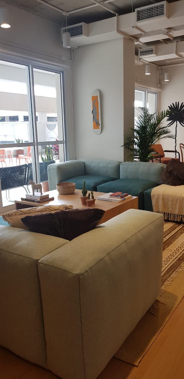 Sala de estar iluminada com sofá bege, poltrona azul, mesa de centro, plantas e janelas amplas.