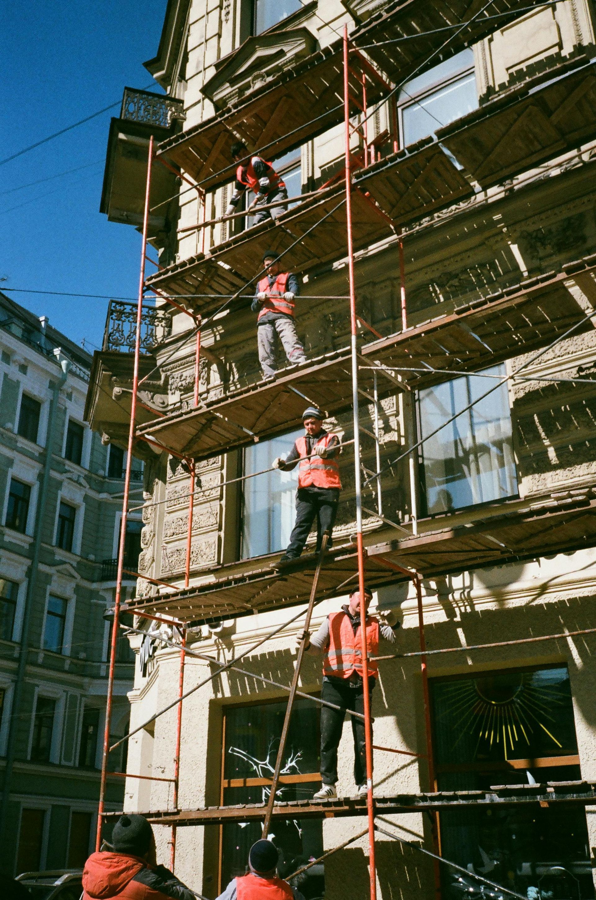 Operários em andaimes ao lado de um prédio em construção, com equipamentos de segurança vermelhos e céu azul.