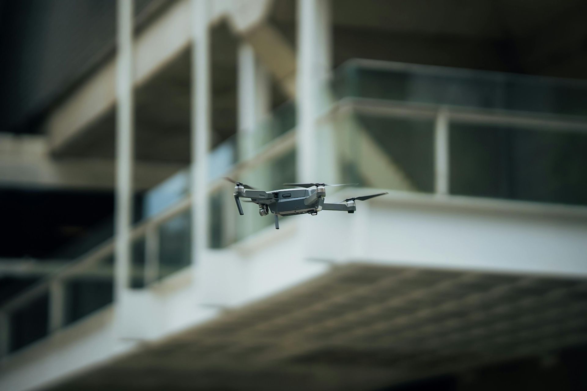 Pequeno drone cinza pairando em frente à varanda de um prédio moderno.
