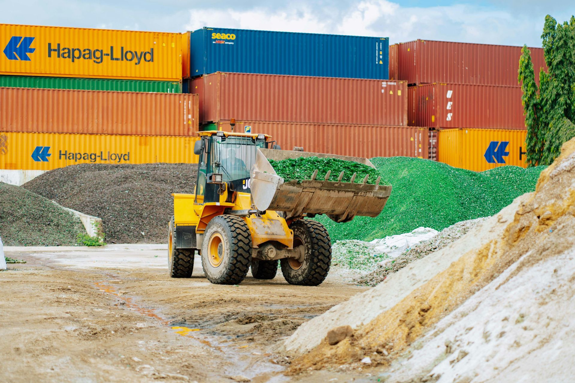 Pá carregadeira transportando cascalho em um pátio de contêineres com contêineres Hapag-Lloyd empilhados.