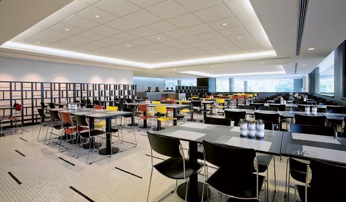 Cafeteria moderna e vazia, com fileiras de mesas e cadeiras sob luzes brilhantes no teto e grandes janelas.