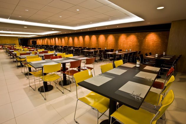 Cafeteria moderna e vazia, com fileiras de cadeiras e mesas amarelas sob iluminação aconchegante.