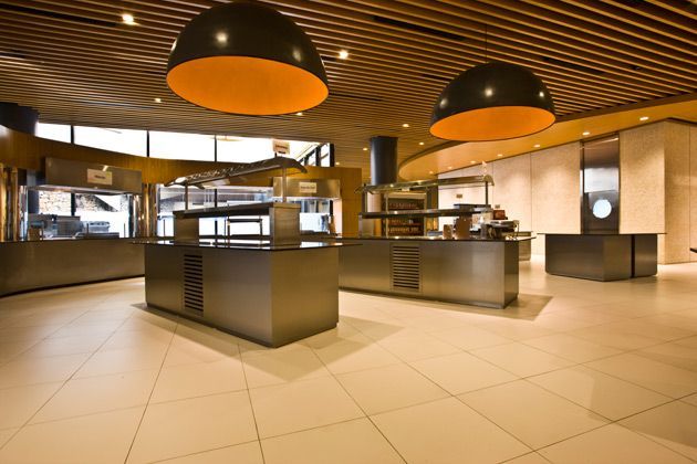 Cafeteria moderna com luminárias pendentes pretas, piso de azulejo e balcões de atendimento em aço inoxidável.