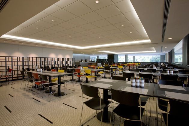 Cafeteria moderna com mesas, bancos e estantes de livros sob luzes brilhantes no teto.