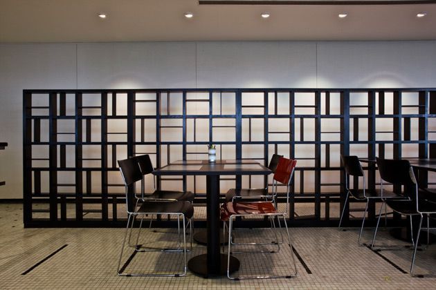 Sala de estar moderna com mesa alta, cadeiras variadas e uma parede divisória quadriculada preta ao fundo.