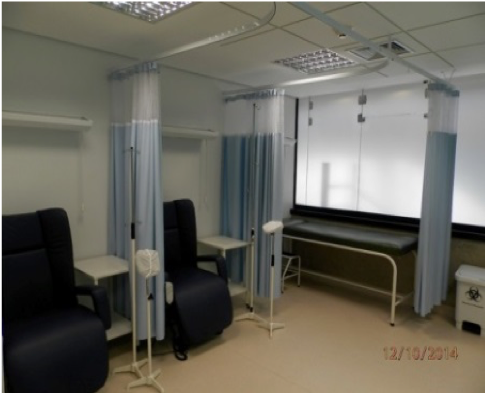 Sala de tratamento hospitalar com cortinas azuis, cadeiras pretas e área de leito com janela.