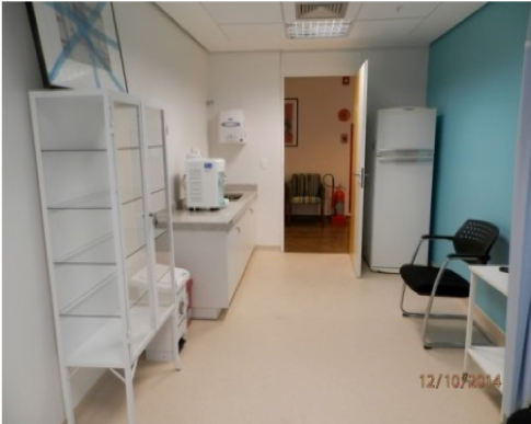 Sala de clínica vazia com prateleiras brancas, mesa de exame e parede com detalhes em azul-petróleo.