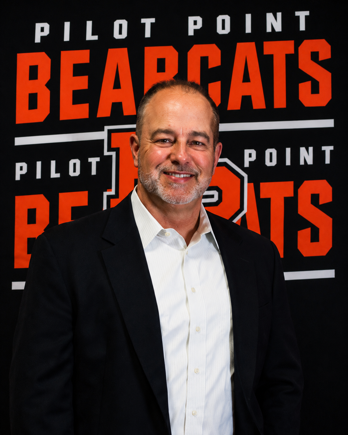 Football coach with arms crossed, standing on a football field. He wears a navy polo shirt and gray pants.