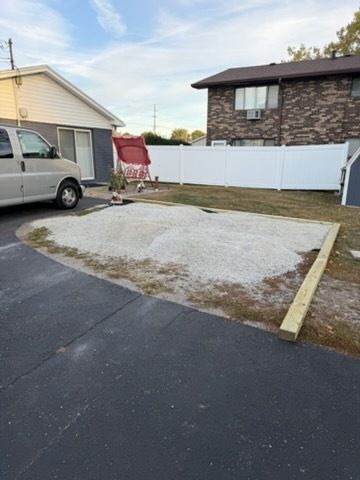 A gravel pad with a wooden border sits between an asphalt driveway, a white fence, and two houses on a sunny day.