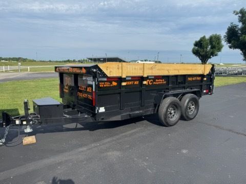 A black dump trailer with wooden side extensions parked on asphalt under a blue sky.