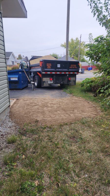 A dark-colored dump truck is parked on a driveway near a blue dumpster, with a large pile of gravel on the ground nearby.