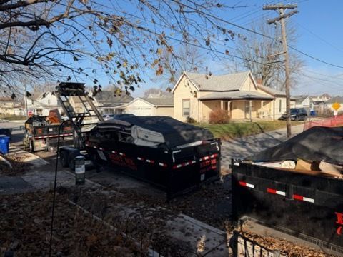 A black roll-off dumpster truck is positioned on a residential street next to another large dumpster in front of a house.