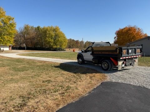 A dump truck sits on a gravel driveway next to a grassy lawn under a clear blue sky, with autumn trees in the background.