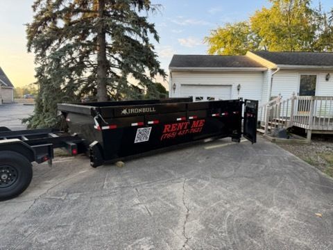 A black roll-off dumpster parked in a residential driveway with open rear doors and a phone number on its side.
