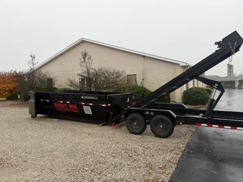 A black roll-off dump trailer sits on a gravel lot next to a building, partially raised by its hydraulic mechanism.