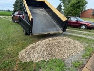 A red pickup truck with a raised dump trailer bed, unloading a pile of gravel onto a grass and gravel driveway.