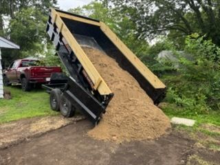 A red pickup truck is dumping a large load of sand from a black trailer onto a residential yard.