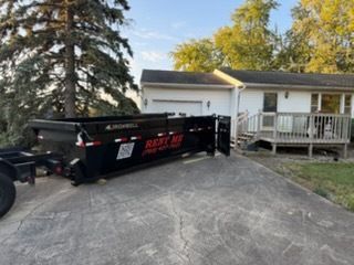 A black dumpster trailer parked on a residential driveway in front of a house with a wooden deck.