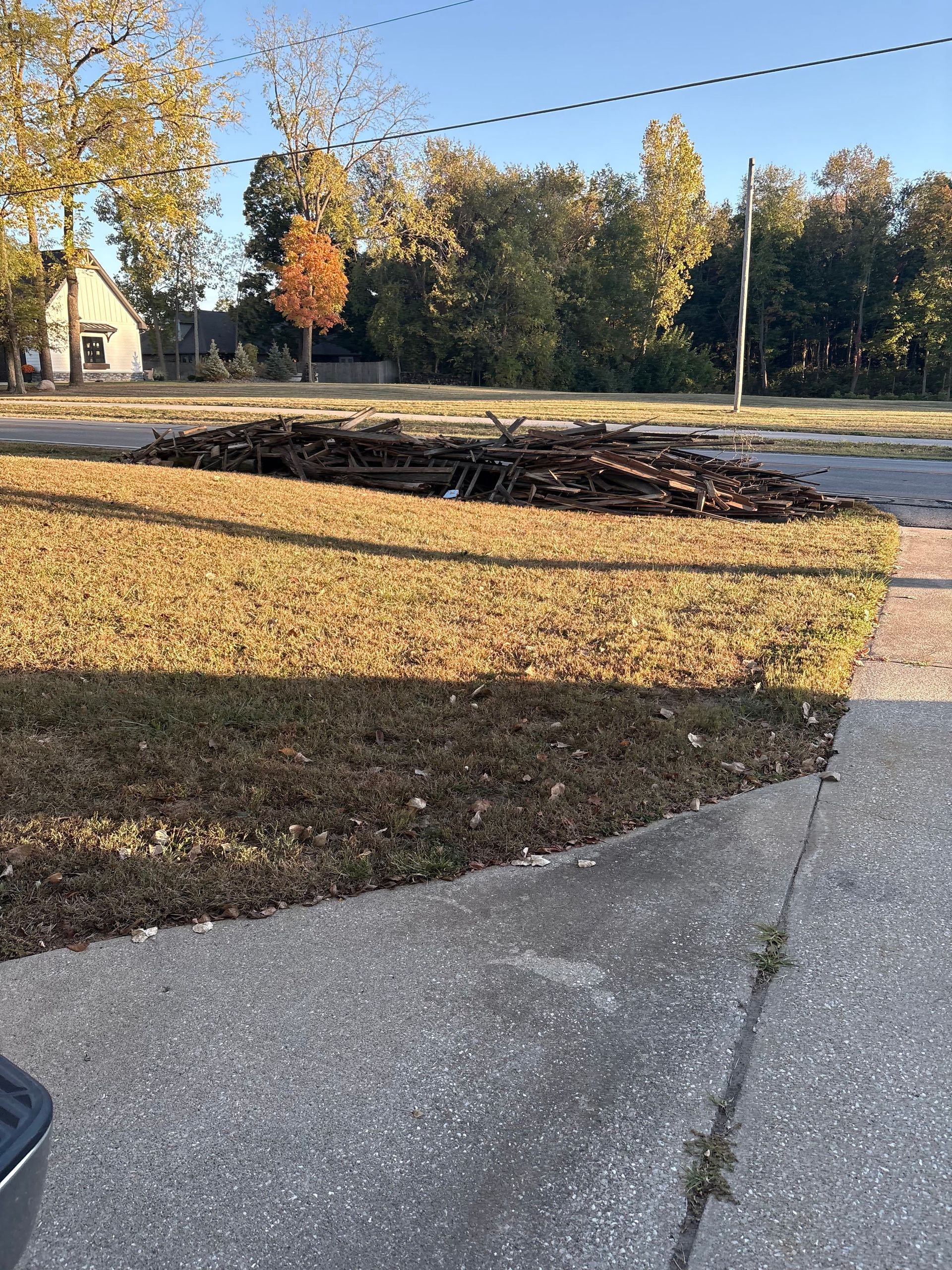 A large pile of dark yard debris sits on the grass next to a concrete sidewalk in a residential neighborhood.