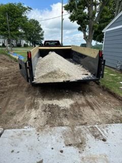 A black dump trailer parked on a dirt lot, filled with a large pile of light-colored crushed gravel or stone.