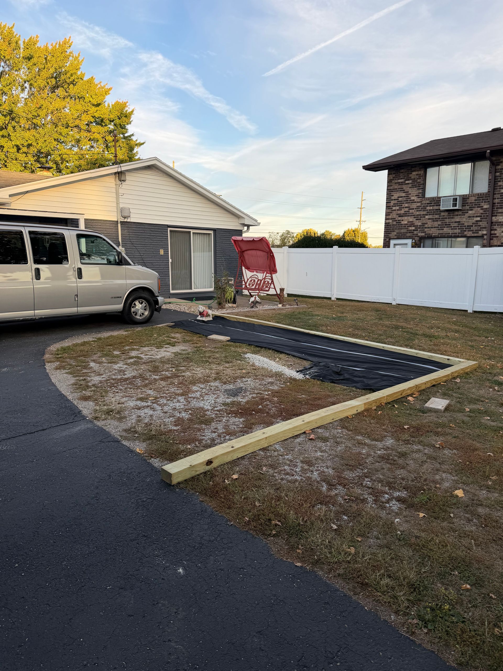 A residential yard under construction with a wooden border, landscape fabric, and gravel near a house and parked van.