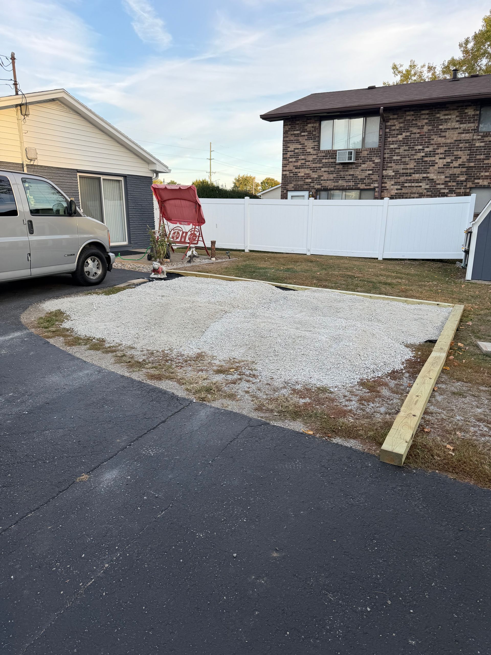A white van parked next to a rectangular gravel driveway pad bordered by wooden beams in a residential yard.