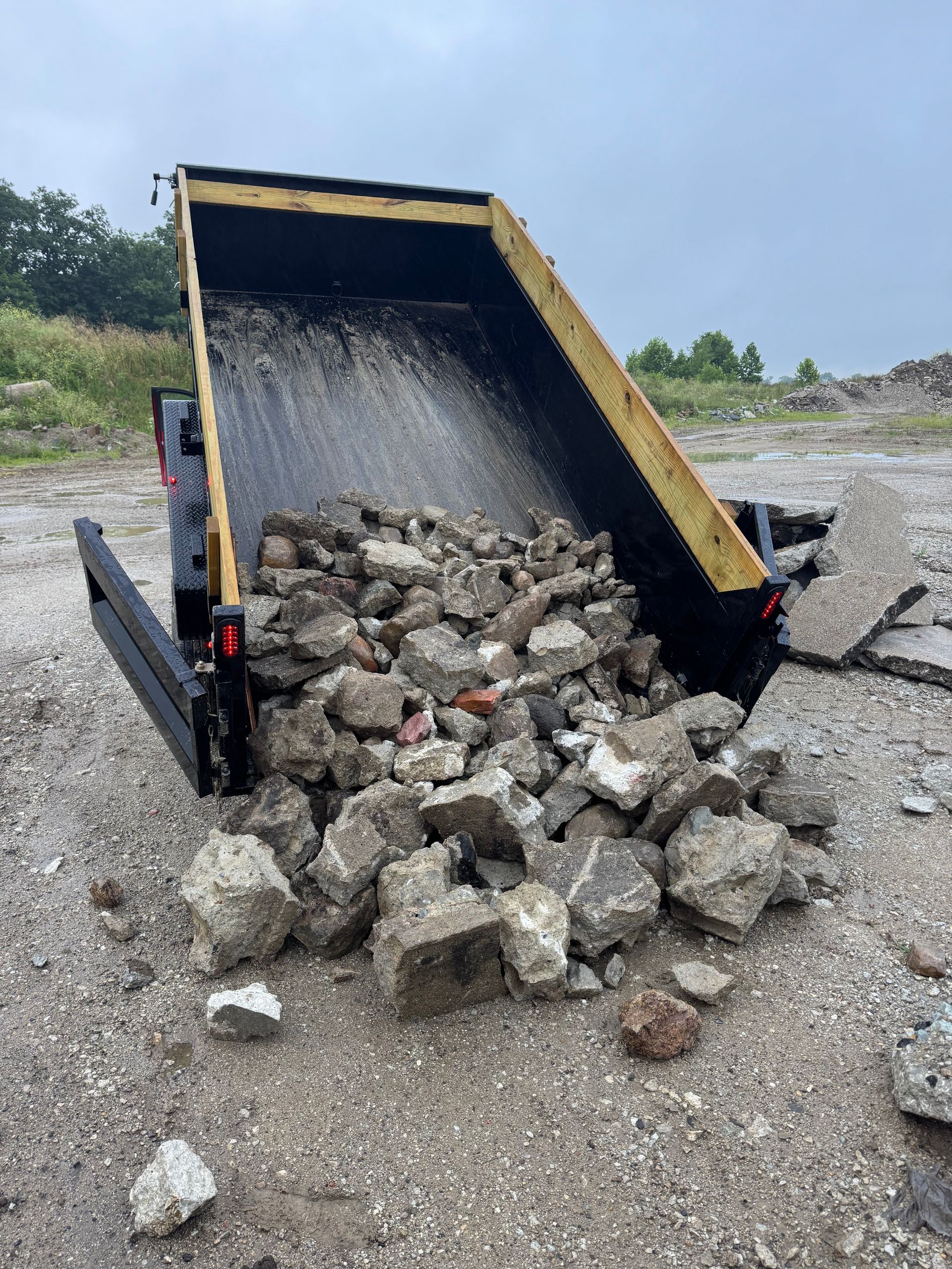 A black dump trailer tipped to the side on a gravel lot, unloading a large pile of broken concrete rubble.