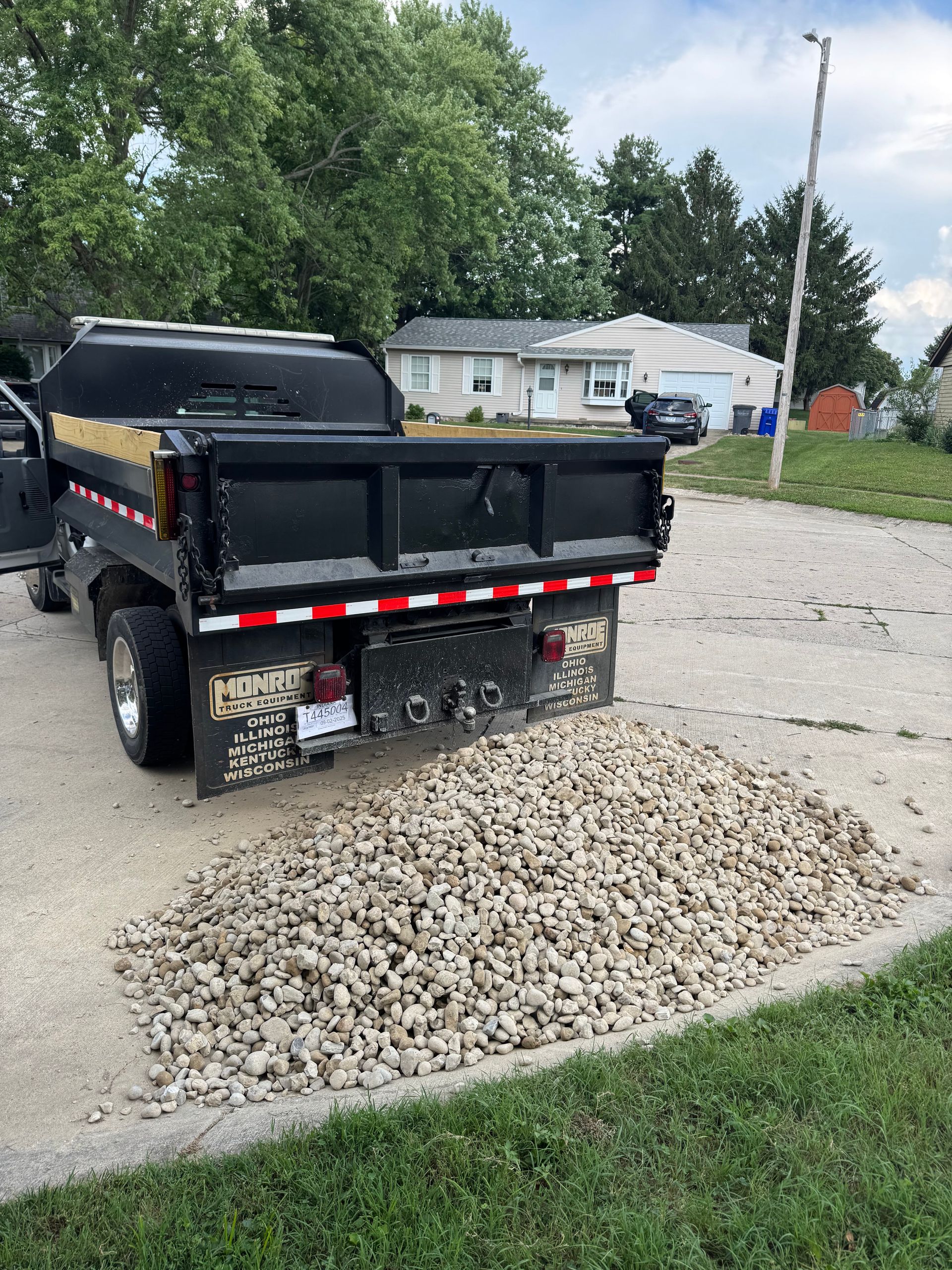 A black dump truck parked on a gravel driveway has deposited a large pile of light-colored crushed rock behind it.
