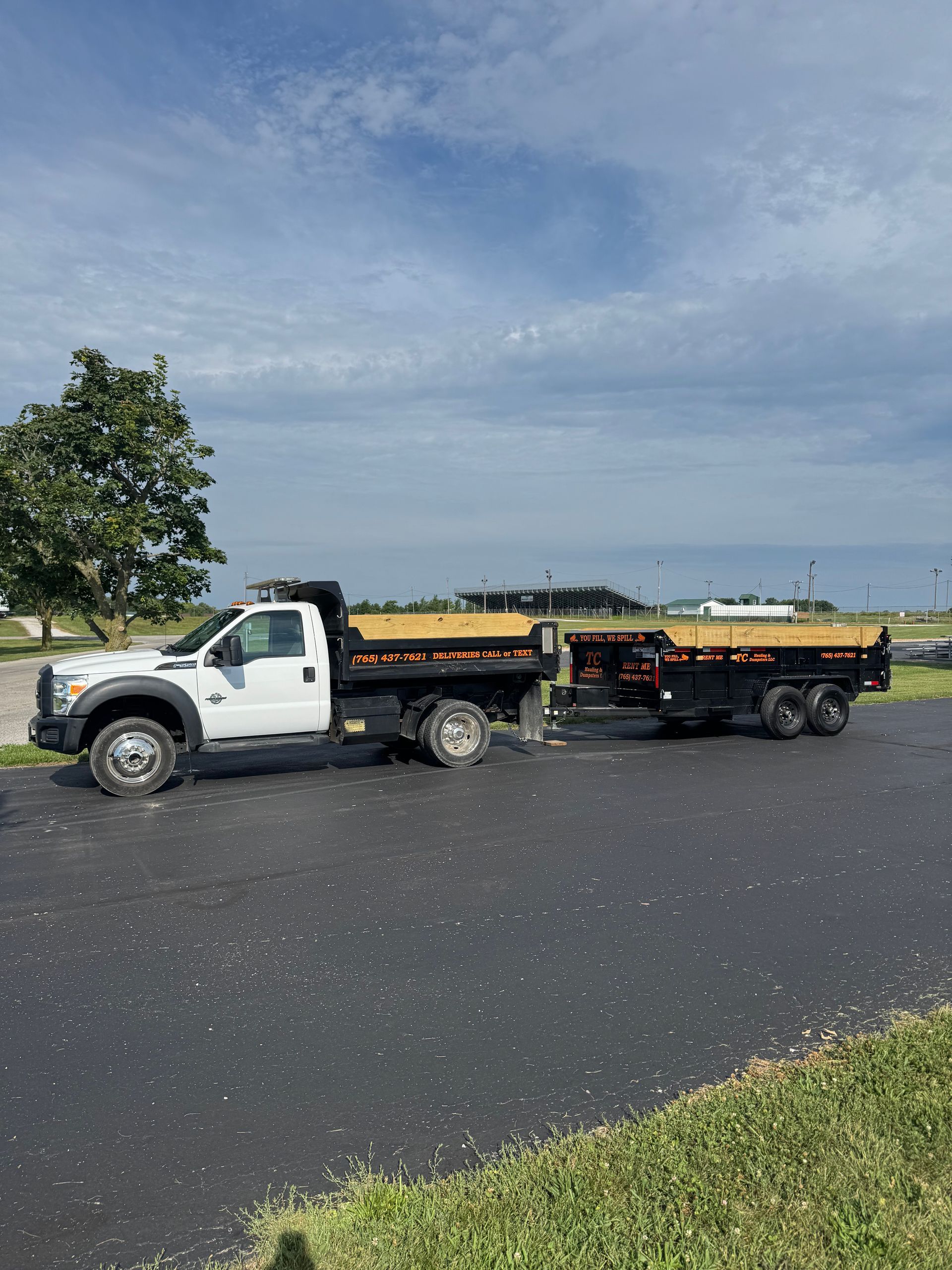 A white dump truck pulling a matching dump trailer, parked on an asphalt lot under a blue sky with scattered clouds.