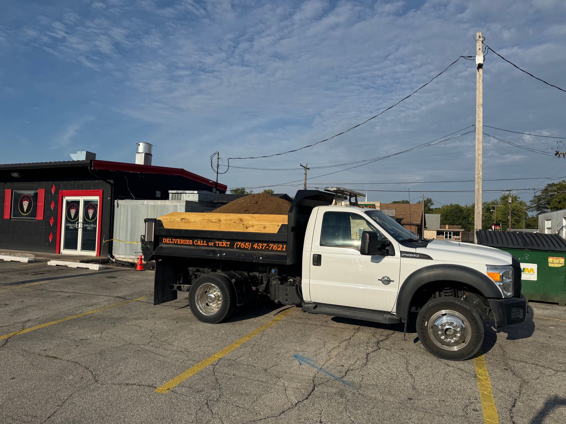 A white dump truck filled with dirt is parked in an asphalt parking lot next to a small, dark-colored commercial building.