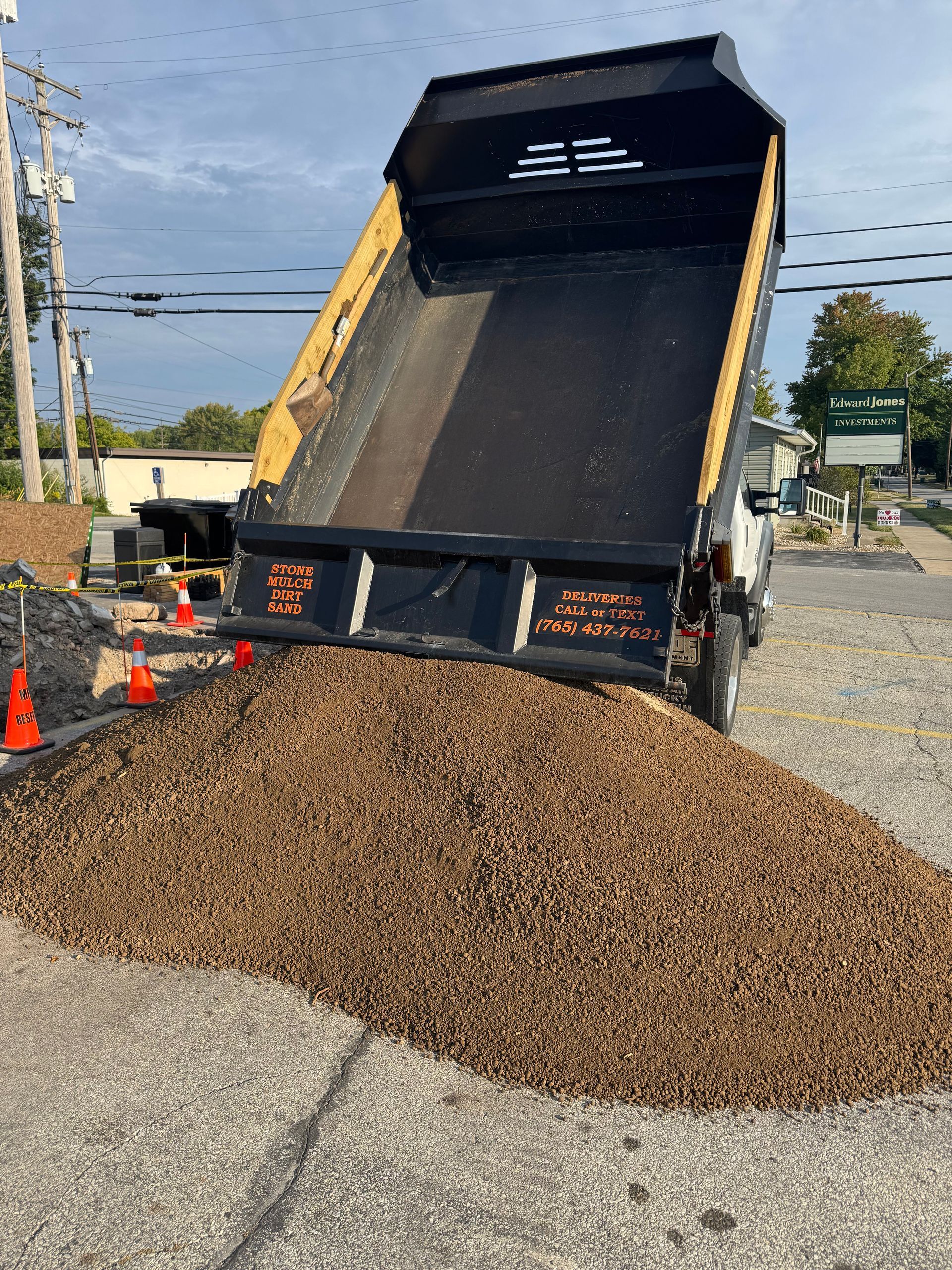 A dump truck lifts its bed to deposit a large pile of brown mulch onto a gravel-covered parking lot.