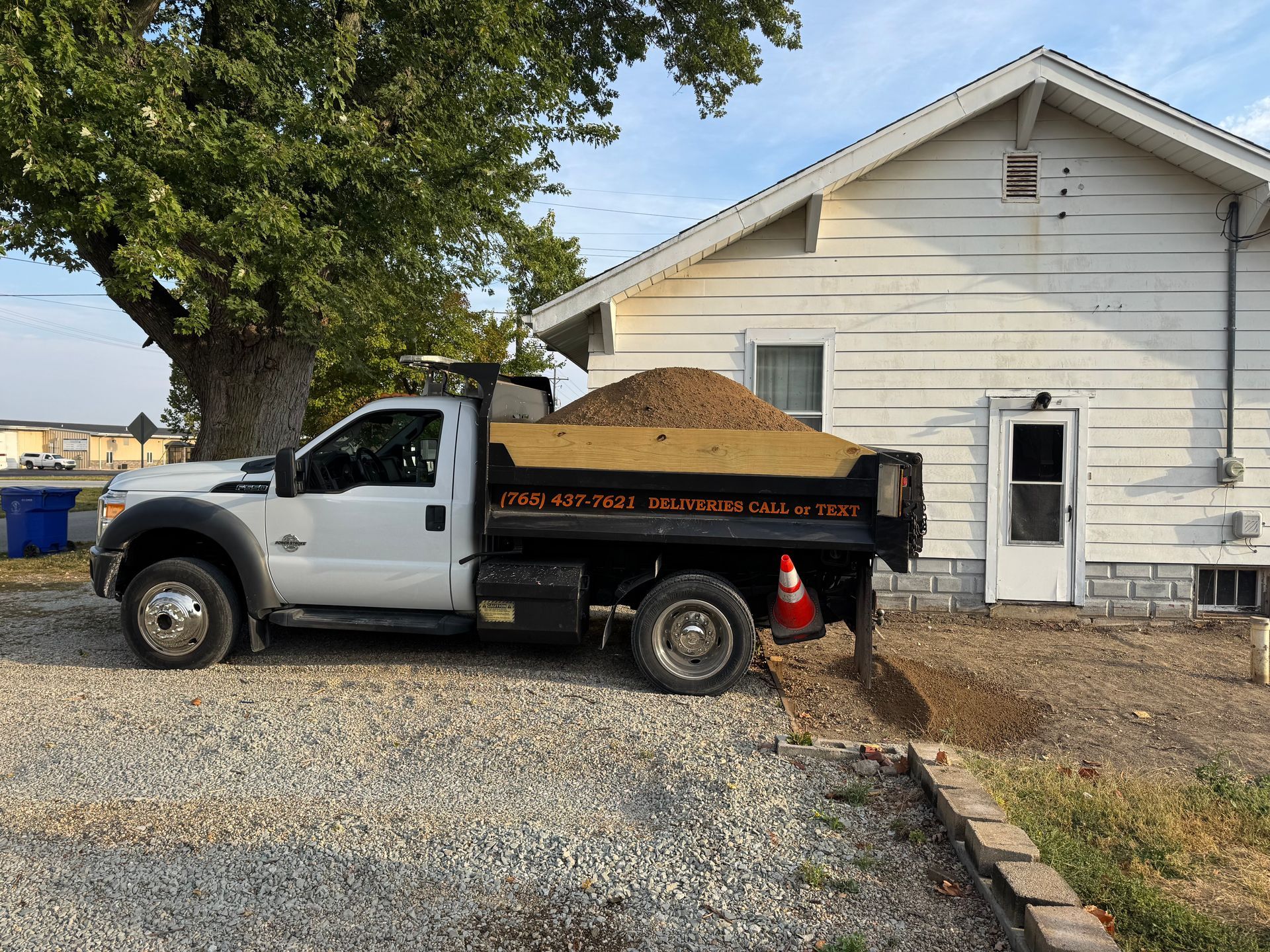 A white dump truck loaded with gravel is parked next to a house, pouring material into a trench along the foundation.