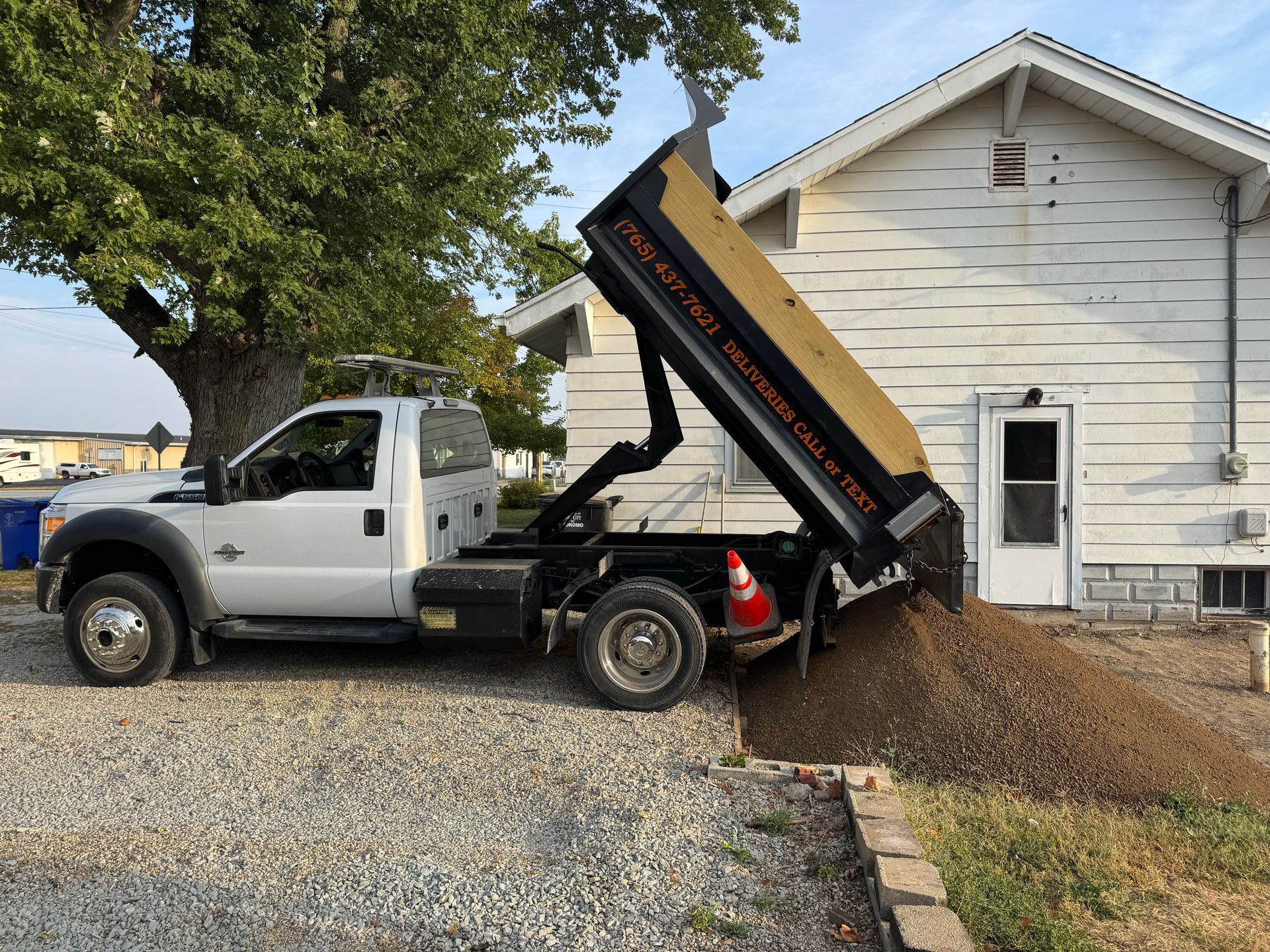 A white dump truck with a raised bed depositing a large pile of gravel next to a white house with grey stone trim.