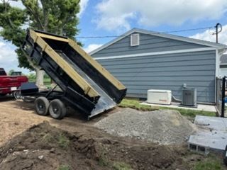 A black dump trailer lifted in an upright position, unloading a pile of gravel next to a blue-sided building.