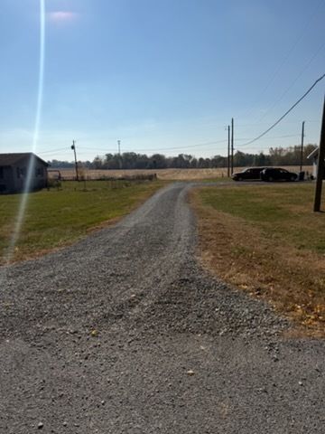 A gravel driveway leads through a grassy yard toward an open field under a sunny blue sky with utility poles visible.
