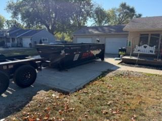 A large black dumpster sits on a residential driveway next to a tan house with a white bench on the porch.