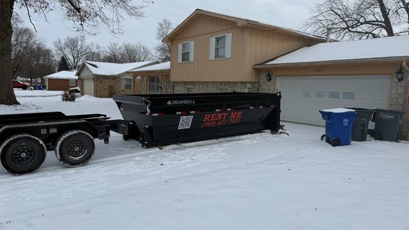 A black dumpster trailer parked on a snowy suburban driveway in front of a tan house.