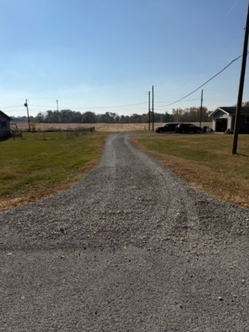 A gravel driveway leads from the foreground toward a field under a clear blue sky, flanked by grassy yards and utility poles.