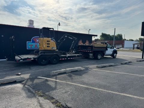 A yellow mini-excavator sits on a trailer attached to a white dump truck in a paved parking lot.