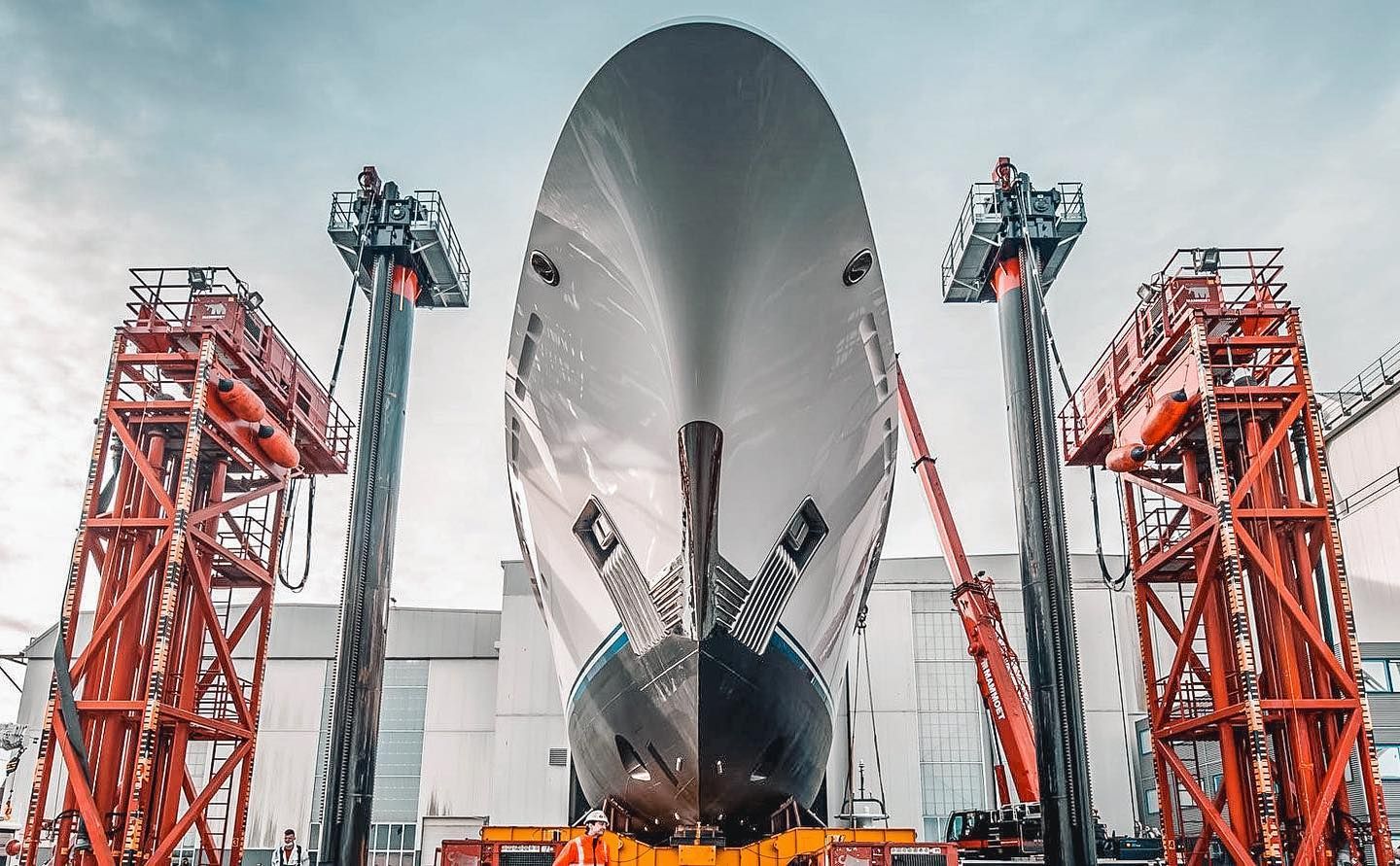 Large ship's bow being worked on by workers in elevated lifts, set against a cloudy sky.
