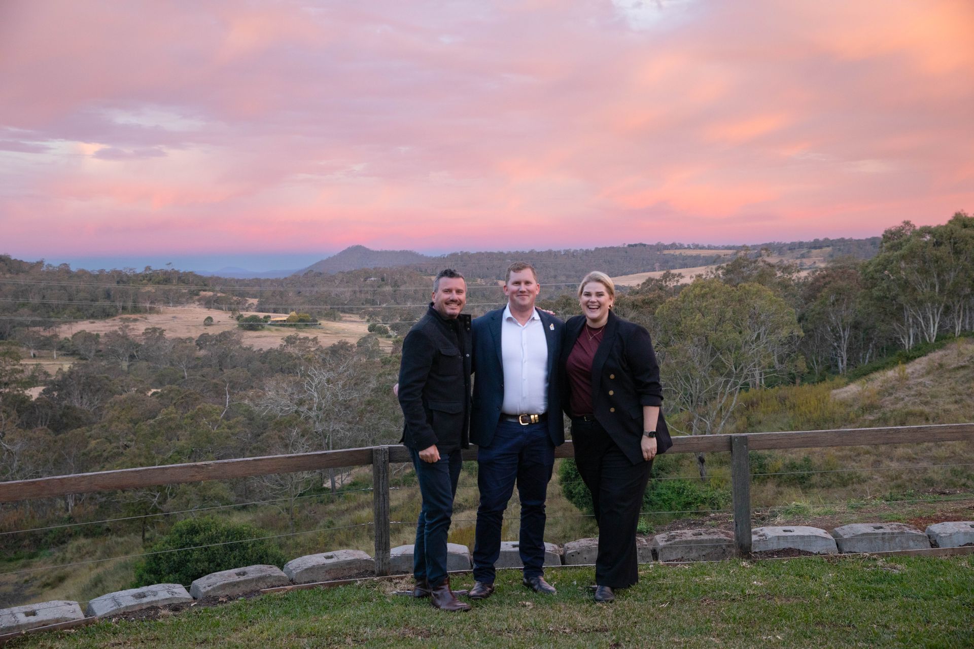 Three people standing on a grassy hill with a sunset in the background. The sky is pink and orange.