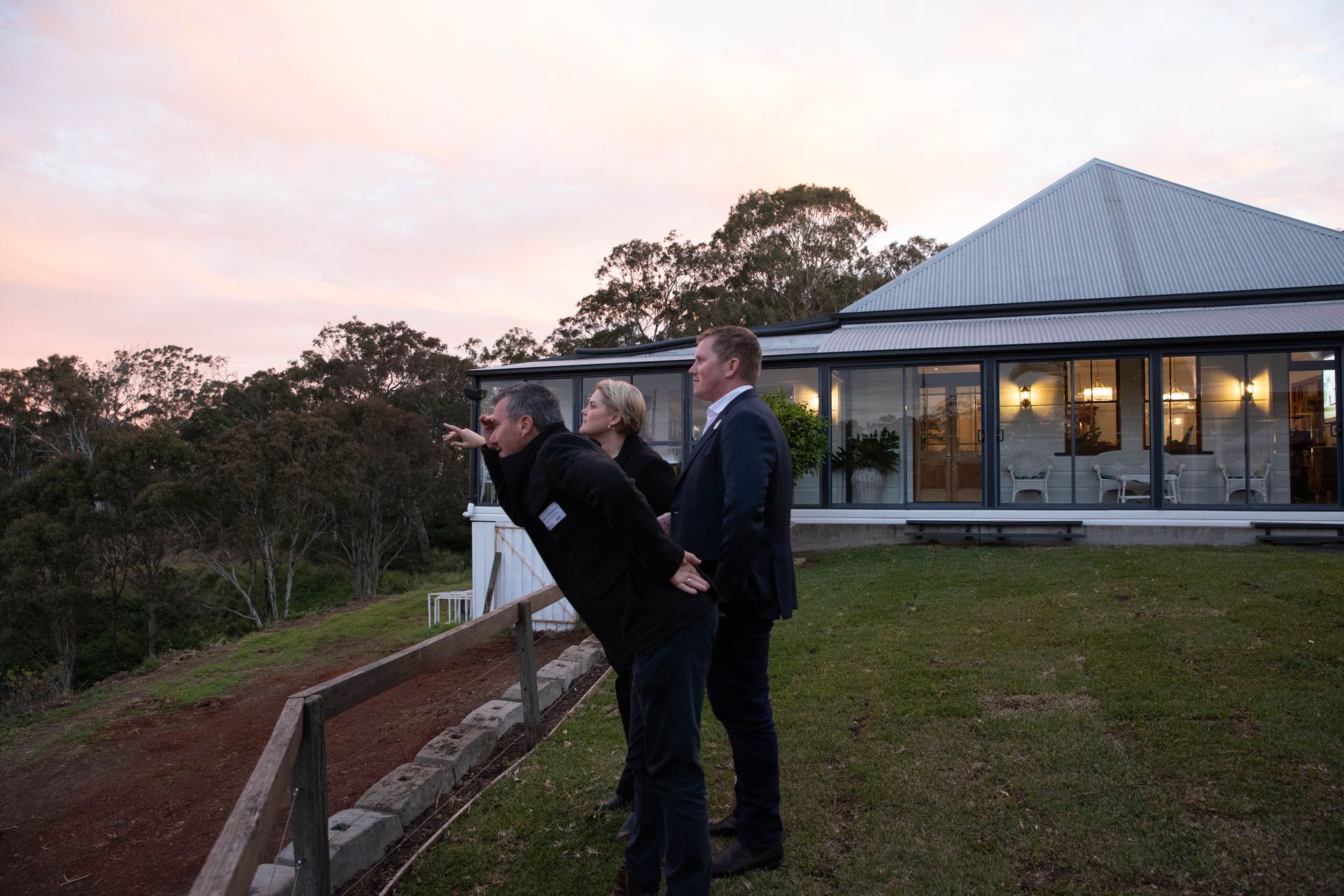 Three people looking at a sunset from a grassy hillside. A house with large windows is in the background.