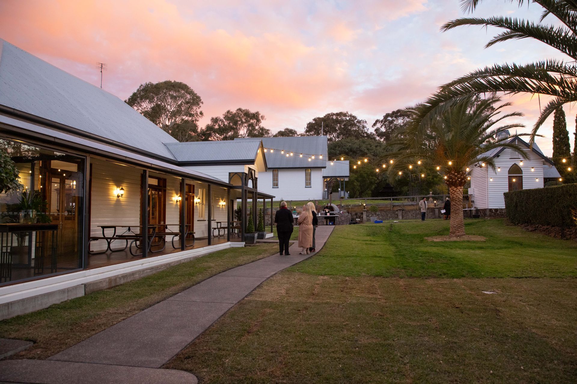 Pathway leading to buildings, two people walking on the path at dusk. Pastel sky and string lights visible.