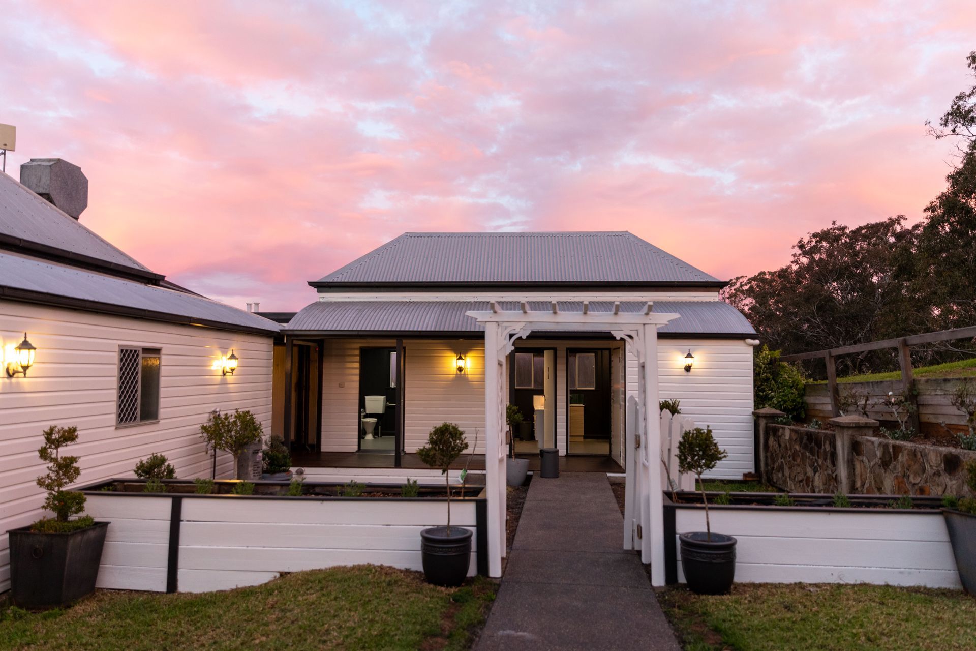 White cottage with grey roof and pergola, illuminated by warm lights against a pastel sunset sky.
