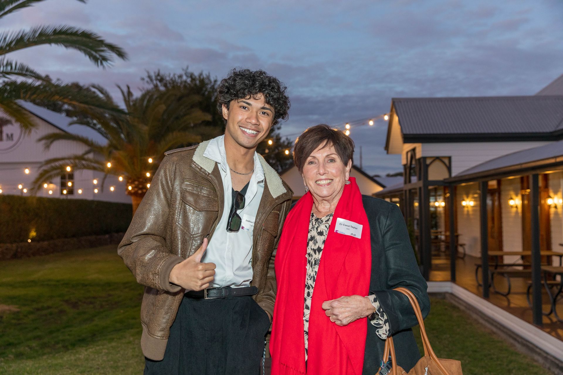 A young man in a brown jacket gives a thumbs-up next to an older woman in a red scarf, outside a white building with string lights.