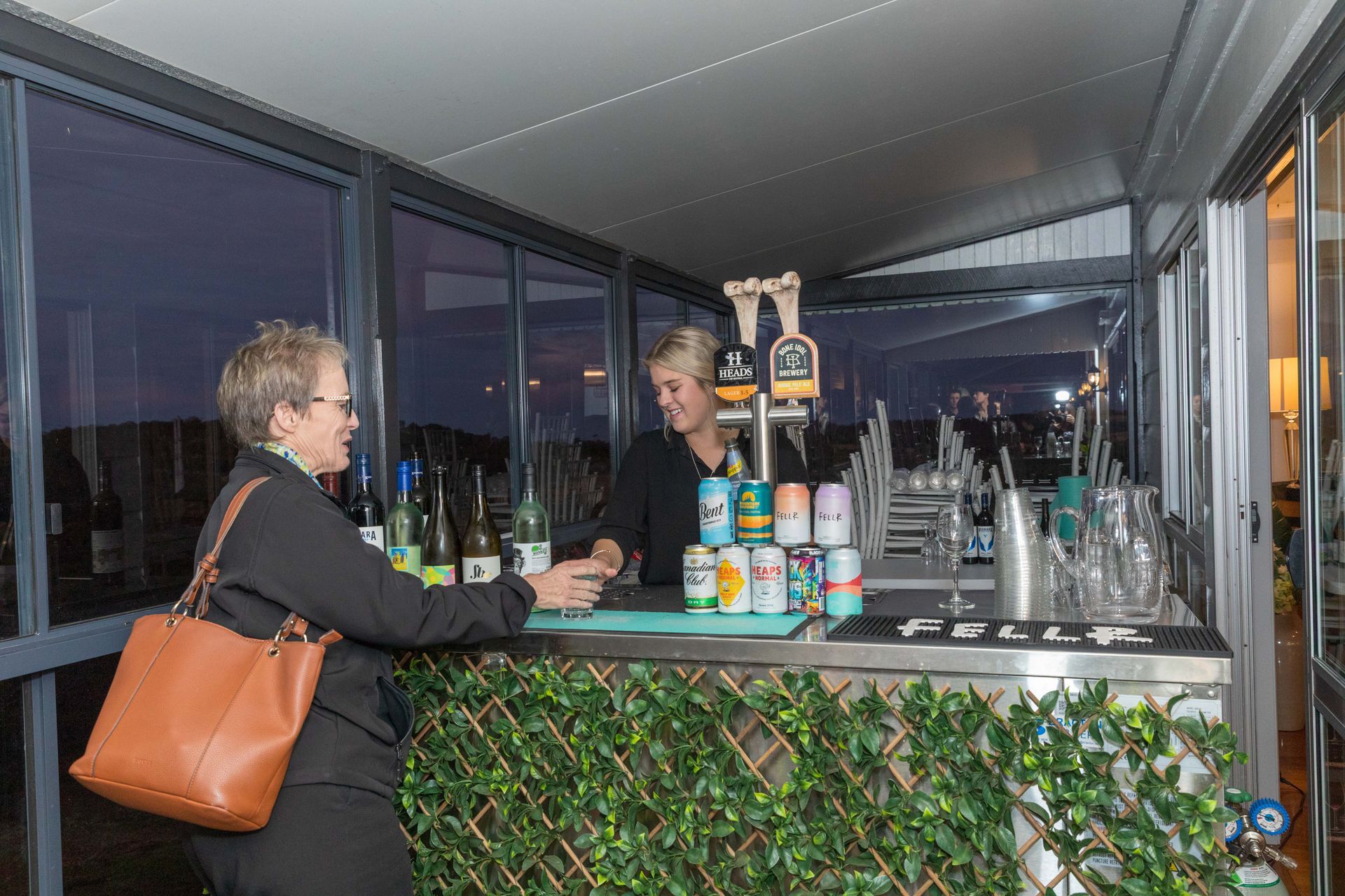 A woman ordering a drink from a bartender at an outdoor bar. The bar is decorated with green vines and features beer taps and canned beverages.