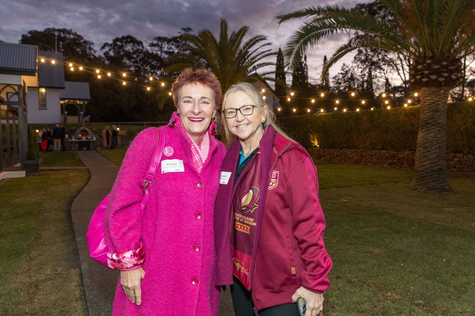 Two women smiling, outdoors. One wears a bright pink coat, the other a burgundy jacket. A building and string lights are in the background.