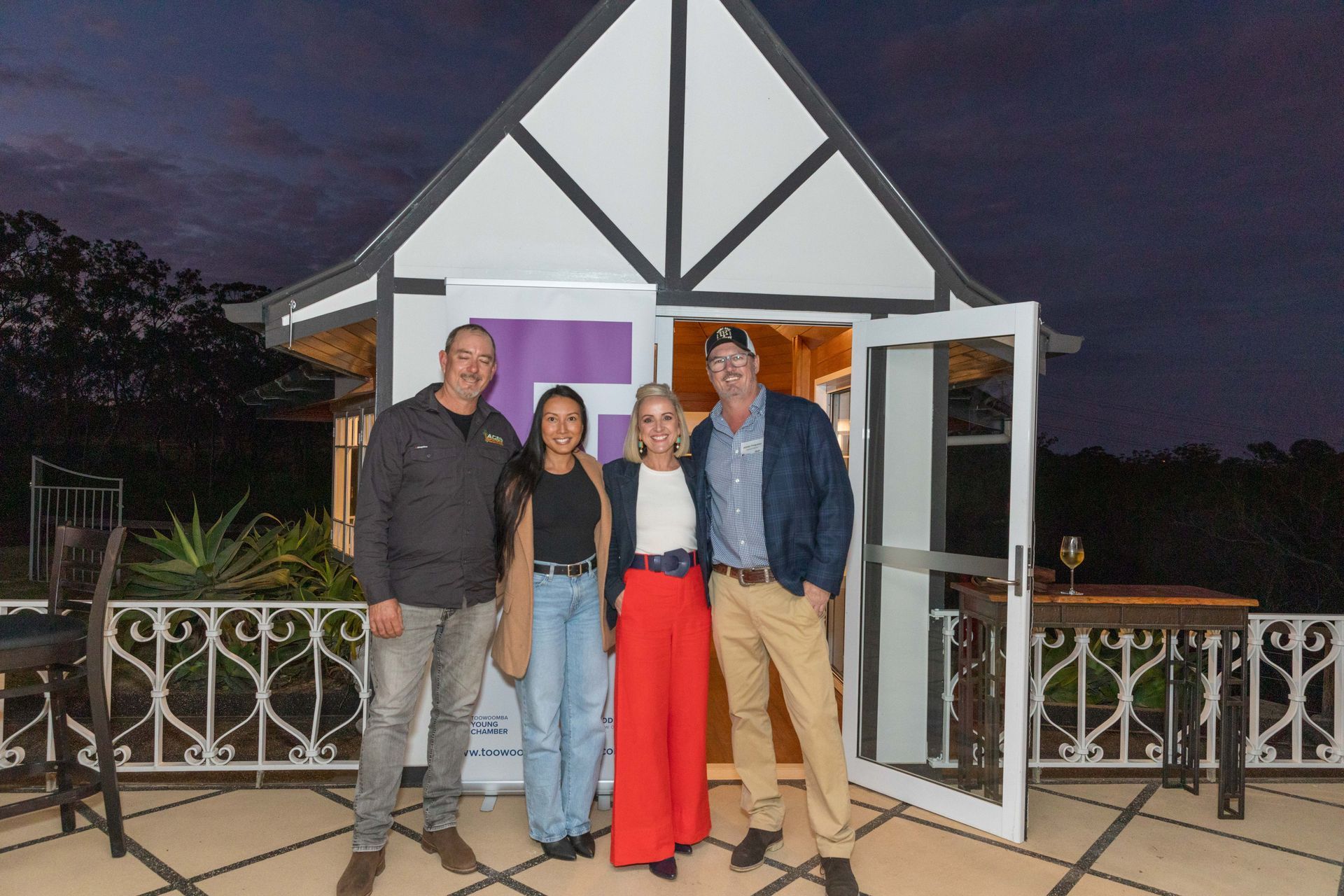 Four people pose in front of a small white and black building with an open door on a patio at dusk.